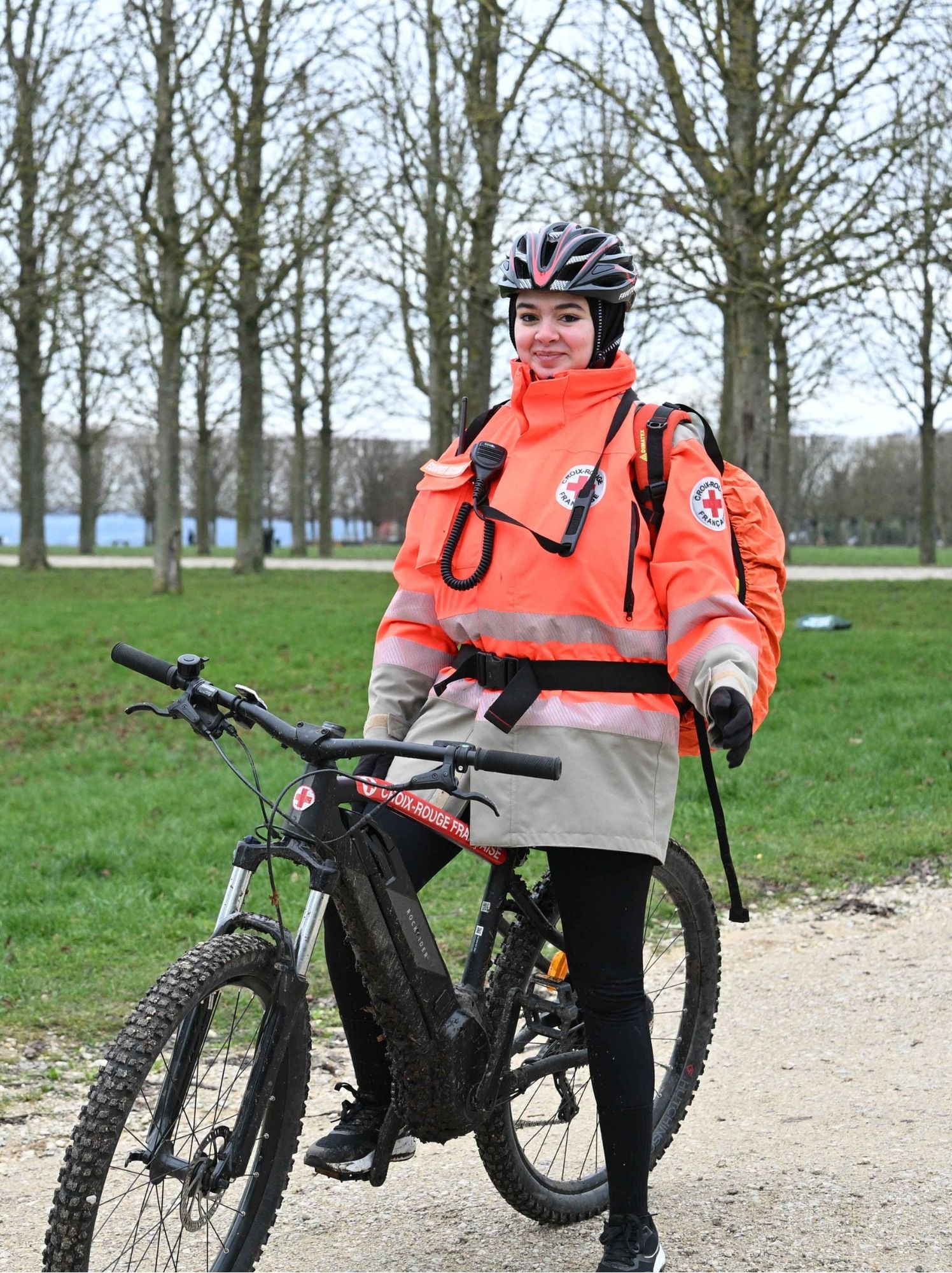 Secouriste de la Croix-Rouge en veste orange fluo, souriante sur son VTT sur un chemin